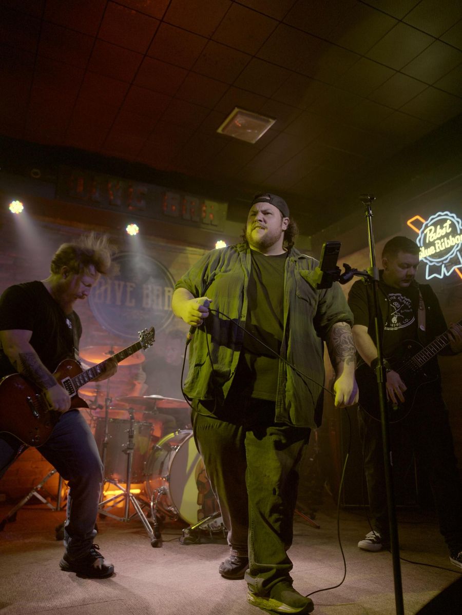 Corey Dow singing centre stage at a dive bar, flanked by both guitarists, with the drummer in the haze behind